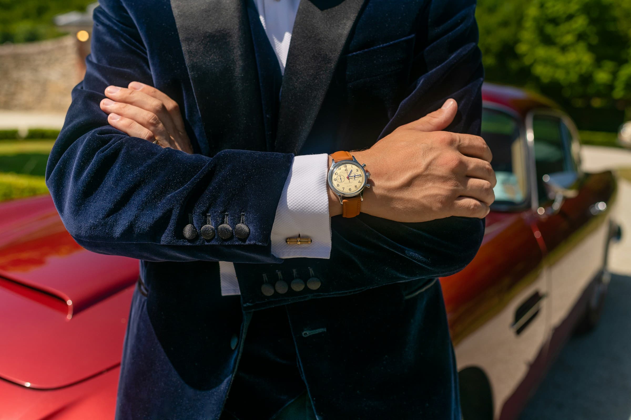 A man in a navy velvet suit, arms folded, wearing a gold watch on a leather strap